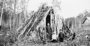 A woman and two children stand before a shelter composed of stacked branches and logs