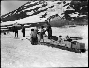 A loaded dogsled sits in the snow with people alongside