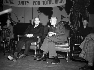 Two men sit with a banner behind them reading "Total unity for total war"