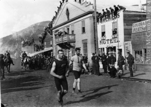 Two men running through a dirt road with a crowd watching from a boardwalk