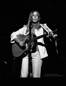Joni Mitchell standing at a microphone with a guitar