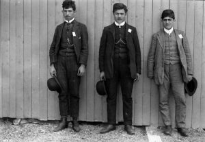 Three suited men stand against a wall, hats in their hands