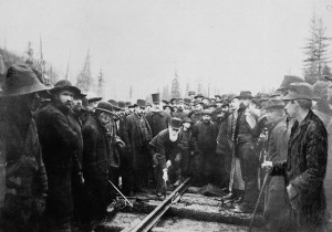 A man hammers a rail spike while a surrounding crowd watches