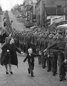 A boy runs to grab the hand of a soldier walking in line through a street