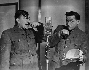 Two men in soldier uniforms speak into a microphone, one holding a comb like a mustache