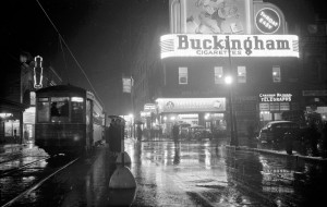 Street with neon signs reading "Buckingham Cigarettes" and "Sweet Corporal Cigarettes"