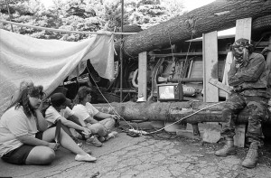 Four people sitting in an encampment composed of an overturned vehicle, logs, and canvas