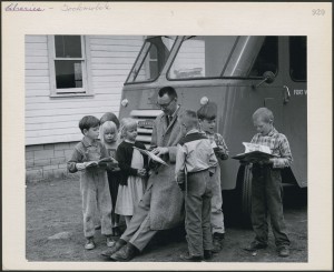 A man and a group of children stand in front of a truck reading books