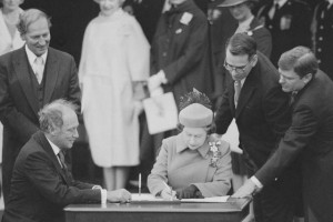 A crowd stands around a table as the queen signs a document