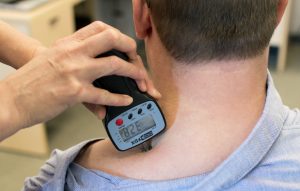 Photograph of a patient (posterior view, neck and head). The neck of the patient's t-shirt is pulled aside, and a clinician is applying an algometer to the intersection of the patient's neck and shoulder