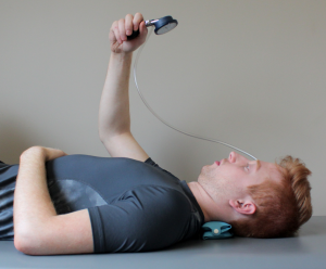 Photograph of a patient lying on their back, shown waist up, lateral view. There is a pressure biofeedback cuff placed under the neck. The patient is looking at a dial held up in their right hand.