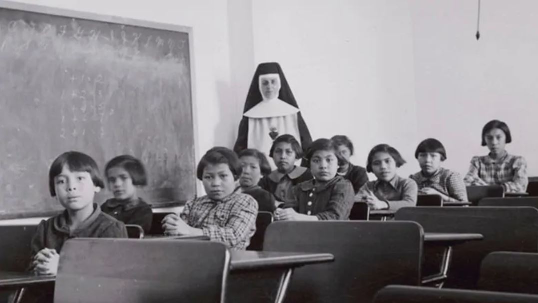 Group of female students [7th from left Josephine Gillis (nee Hamilton), 8th from left Eleanor Halcrow (nee Ross)] and a nun (Sister Antoine) in a classroom at Cross Lake Indian Residential School, Cross Lake, Manitoba, February 1940. Credits to: Canada Dept. Indian and Northern Affairs/ Reuters / Library and Archives Canada