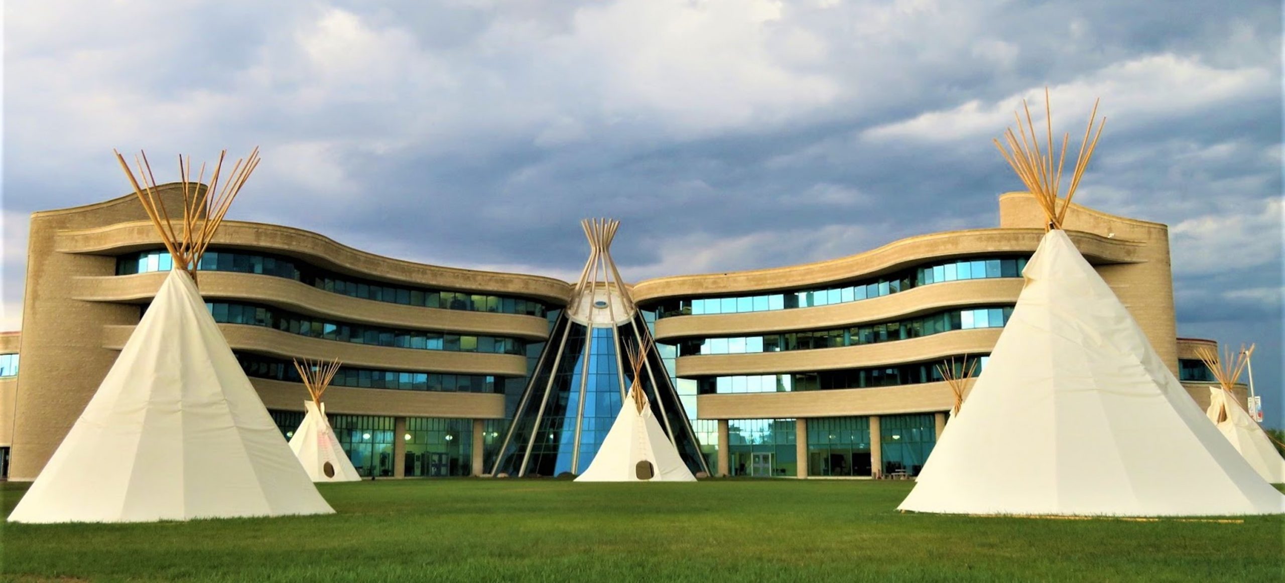 Several tipis laid out symmetrically in a circle on a lawn, with the main building of First Nations University at least 100 metres away in the background. The main building is about five times as wide as high, and is undulating, with half-storeys of glass windows alternating with half-storeys of sandy-coloured concrete. The main floor is entirely glass with rounded concrete pillars every twenty feet or so. The main entrance is a large glass half-tipi which is taller than the building itself.