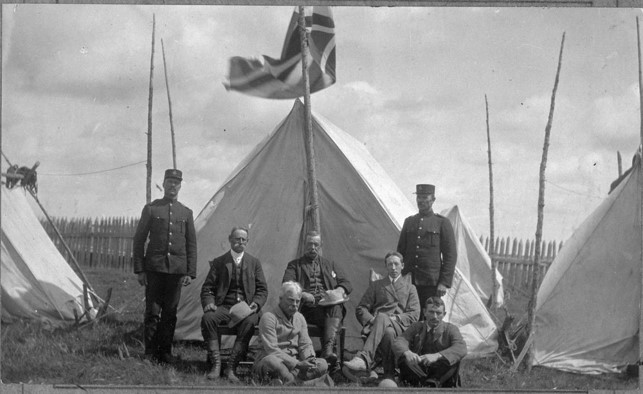 This photo shows a group of seven white men in front of an A-frame canvas tent. A flag of some kind is flying above and behind them, directly in front of the tent, borne on a pole made from a young tree. There are tents on each side of the central tent. Each tent is supported with pegs made from sticks, poles, and ropes tied to poles stuck in the ground outside the poles. There is a tall picket fence in the back ground. Some men are seated on chairs, some are seated on the grass, and some are standing. Two of those standing are dressed in military uniform including a cap. The other men are hatless and dressed in suits. The sky is cloudy.