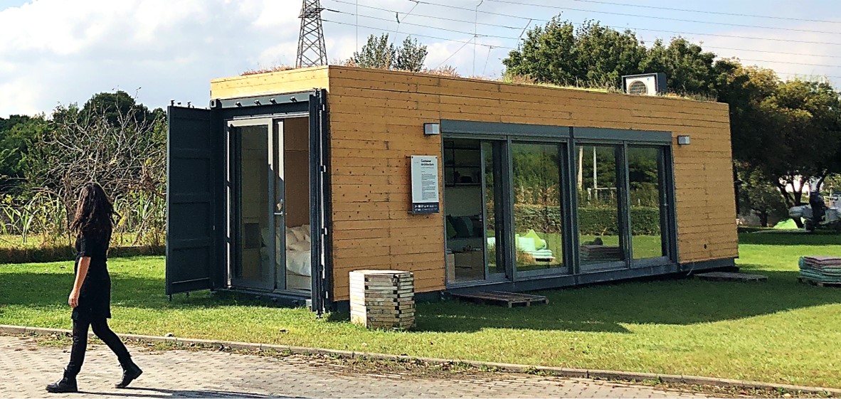 The shipping container has been clad in short planks of oak-coloured wood siding. At the one end of the container which is visible is a sliding glass door, with a protective black metal outdoor door for use when needed. This sliding door takes up most of the width of the end of the container. About 60% of one side of the container features two more sliding glass doors side-by-side, clad in black metal.