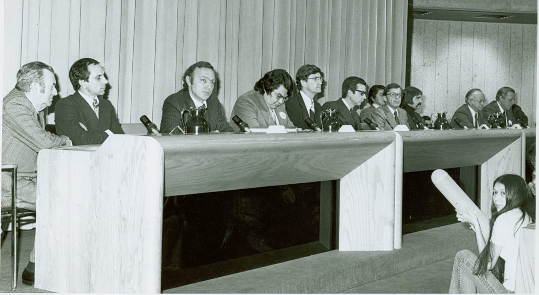 Eleven men in suits at a very long disk below which a female reporter is crouched, holding a giant cylinder, presumably a microphone.