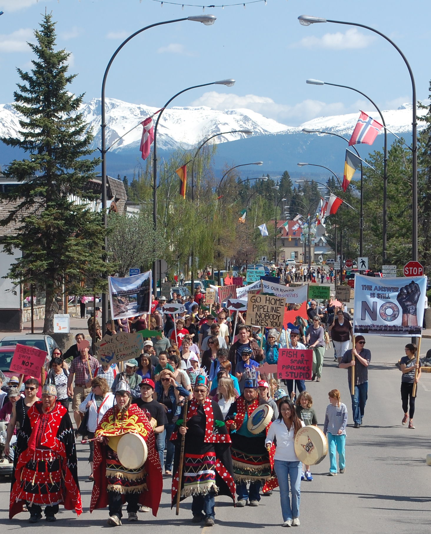 This photo features snow-capped mountains in the distance. A colourful crowd is cheerful marching up a street lined with streetlamps and conifers. Some of the signs being carried read "The answer is still NO" "Go home pipeline, nobody likes you" and "fish are smart, pipelines are stupid". The parade is led by people wearing regalia and people playing drums.