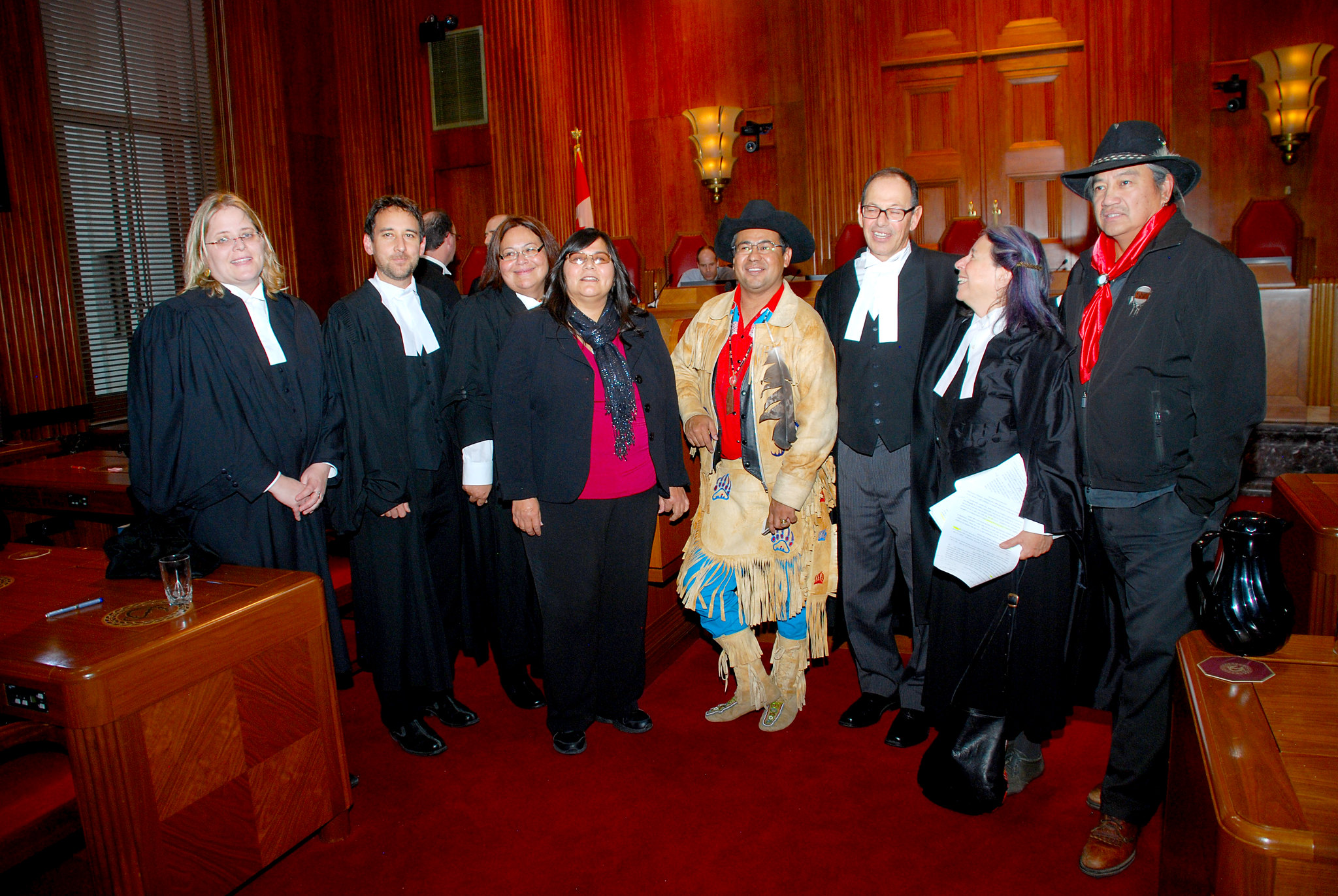 A group of lawyers in formal attire, along with three Indigenous leaders, two of whom are in traditional clothing. The setting is a wood-paneled courtroom.