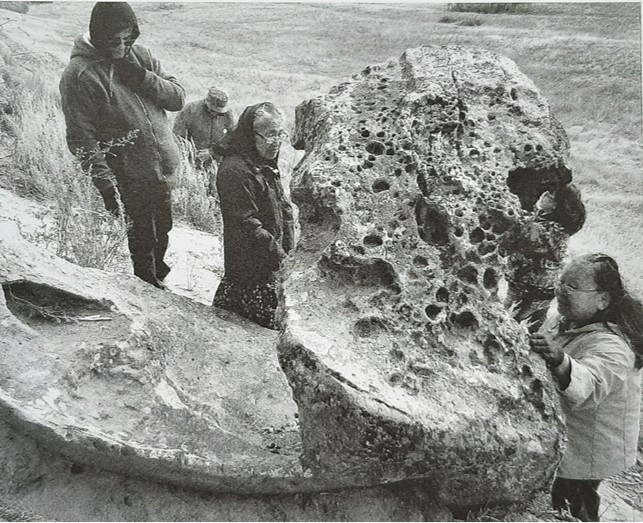 This photo shows four Indigenous elders near the a large, apparently igneous rock formation known as "Coyote's Sweathouse". Three of the elders are near the rock. One is touching it and looking contemplative. Another is staring downward towards it, looking dejected. The third has one of his hands on his cheek and is looking at the ground, projecting sadness.