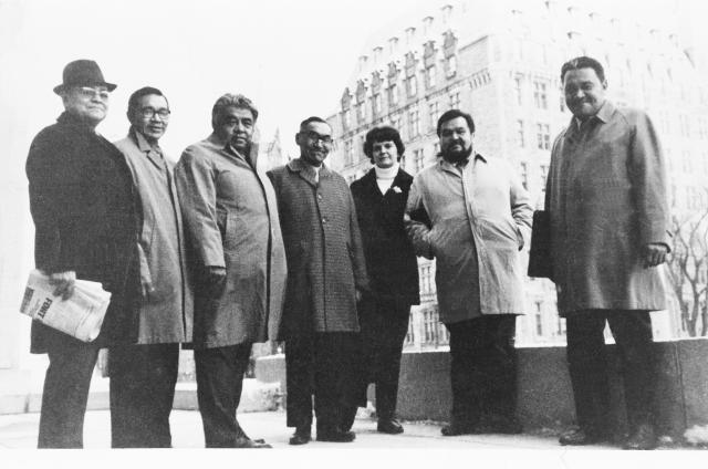 A group of six men and one woman, dressed for the outdoors in cool weather, in front of a tall brick building of the nineteenth century. Most appear to be Indigenous. One is holding a briefcase and Frank Calder, at left, is holding a newspaper. Dr. Calder is the only one wearing a hat.