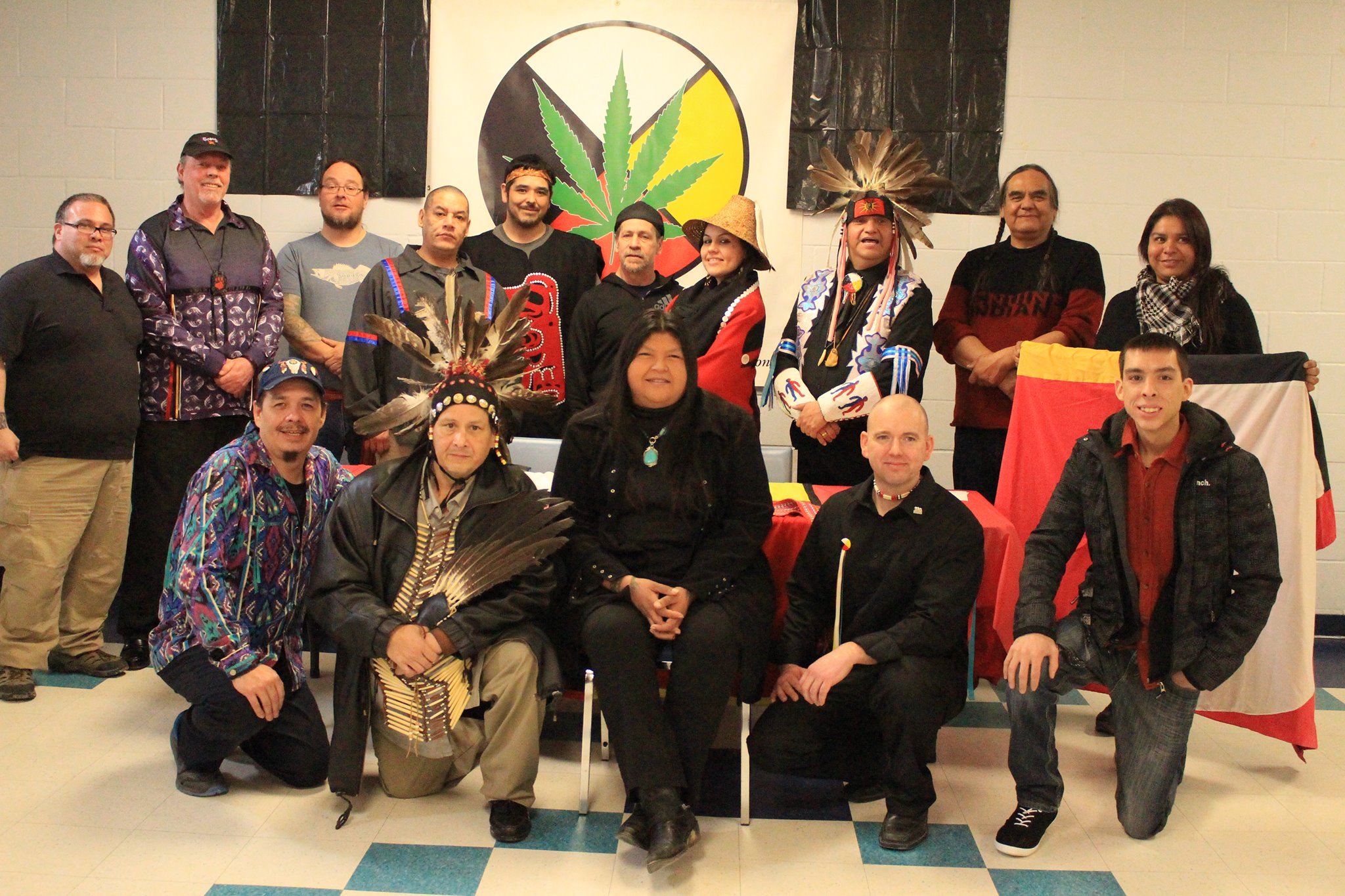 This photo shows a group of fifteen individuals, mostly men, several of whom are in traditional Indigenous dress or regalia from more than one different cultural group. On the wall behind the group is a large medicine wheel divided into four coloured quadrants (black, white, yellow, red). There is a large cannabis leaf superimposed on the medicine wheel.