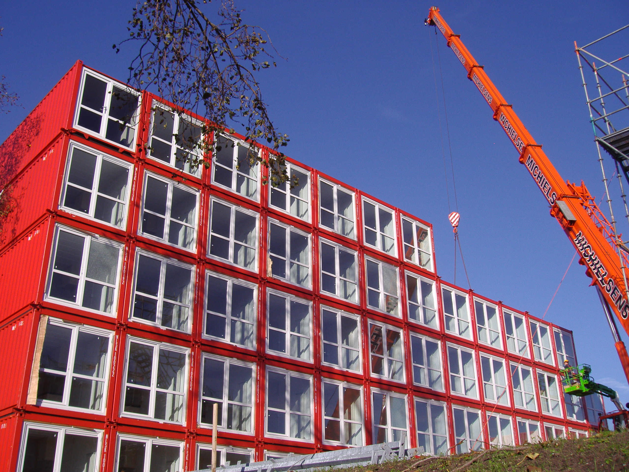 This photo shows a stack of shipping containers, butt ends facing the viewer, five containers high and more than ten containers wide on the base of the building. The shipping containers are red but the butt ends are entirely filled with a window divided in four panes and trimmed in white.