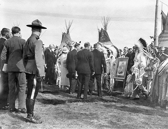 In this picture there is a crowd gathered. There is an RCMP officer, several men in suits, a woman in formal wear, and at least five chiefs in regalia. There are three tipis in the background. There is framed picture on an easel.