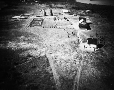 This photograph is blurry. It shows a large field criss-crossed with a few walking paths and roads. There are two or three white frame buildings that might be a church, post office, schoolhouse, a fenced garden, and some oddly shaped barns. There don't seem to be any residential structures for the Indigenous community.