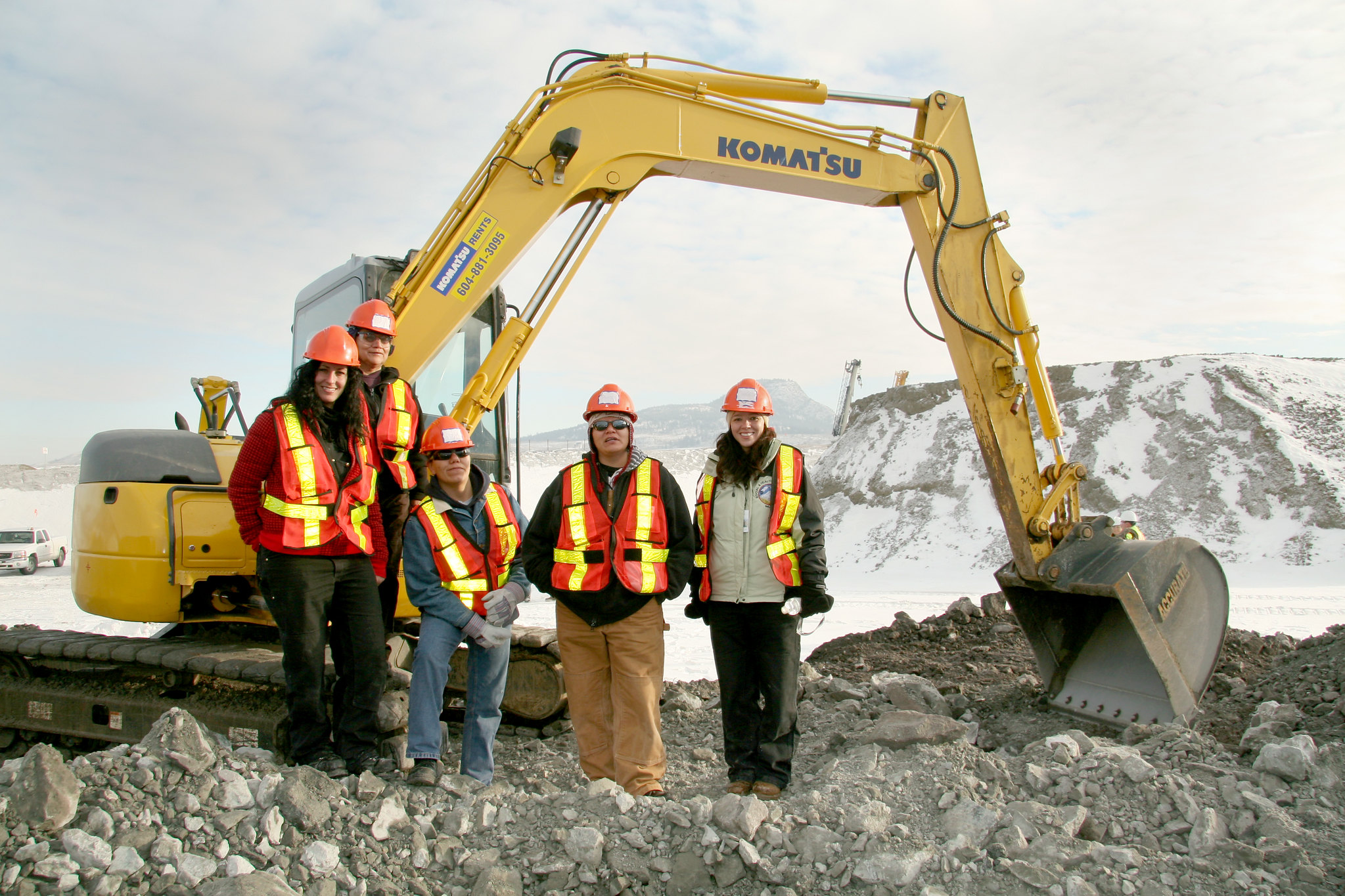 This photo shows five people in hard hats and safety vests in front of a front-end loader on a mining site in snowy landscape.