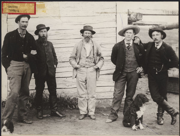 A group of white men in semi-formal attire, striking a relaxed pose at the side of a whitewashed building, with a dog.