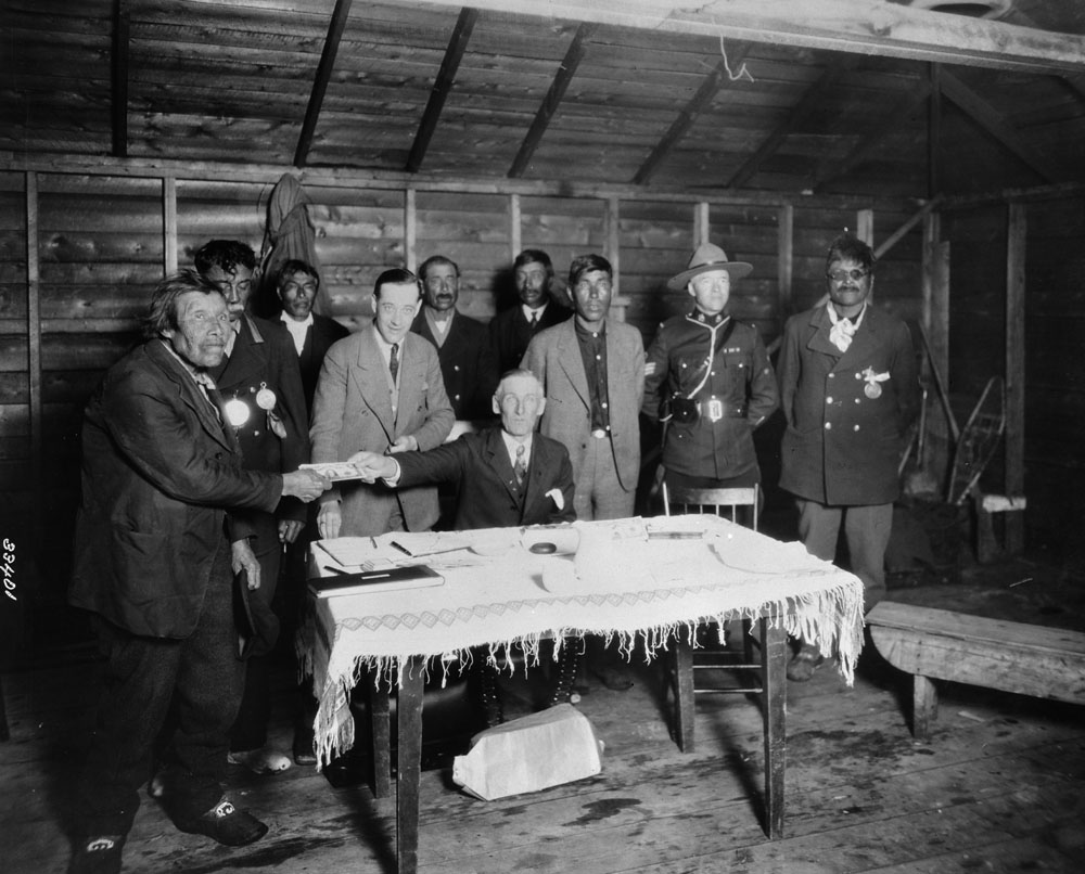 This photo shows a group of people behind a table in a wooden frame building. A white man is seated at the table, which has a woven tablecloth and supports several documents. The man is holding a piece or wad of paper money out to an Indigenous man who is holding the other end of it. There are six other Indigenous men around the table, and two white men, one of whom is a member of the Royal Canadian Mounted Police (RCMP). Everyone is wearing a suit except for the RCMP officer, who is wearing a uniform.