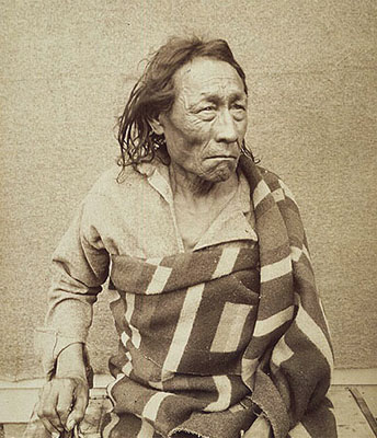 Big Bear is looking away from the camera in a determined way. He is thin and his face is lined. He is wearing a collared shirt which is unsecured at the top. Over that he is wearing a patterned blanket wrapped around his torso with its end coming from behind draped over his shoulder.