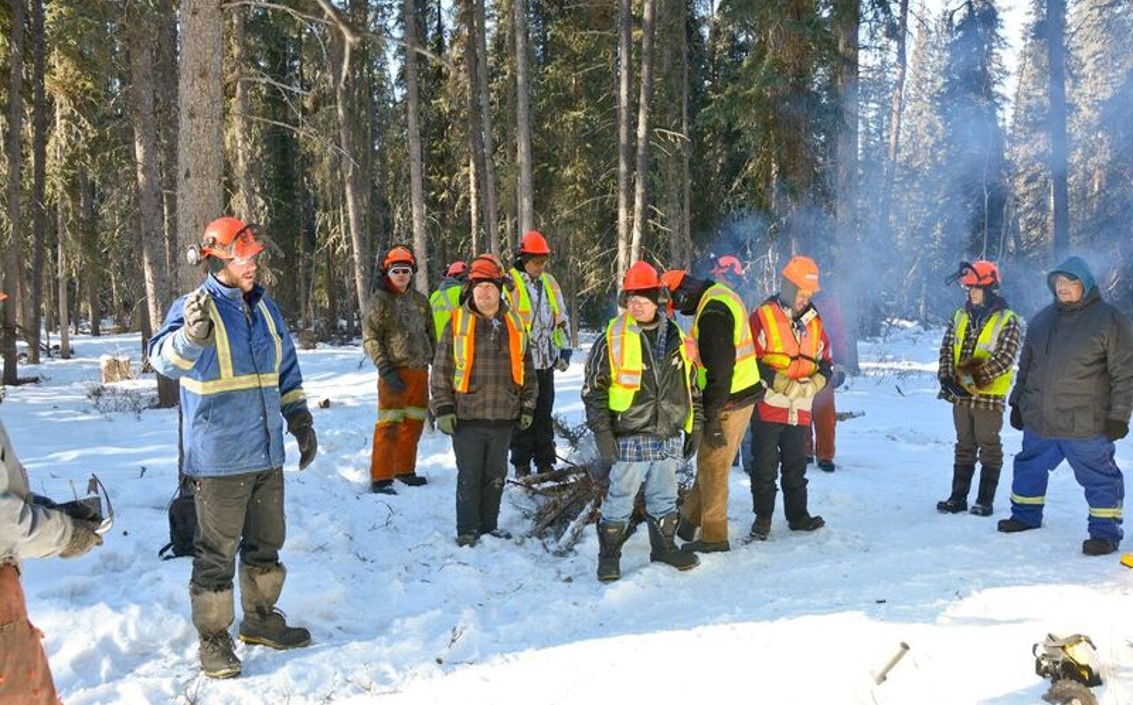 A group of people, mostly men, wearing hard hats, parkas and boots, standing in a clearing in a coniferous forest. There is snow on the ground. There is a small bonfire in the middle of group, mostly obscured from sight, but the smoke can be seen arising to the right.