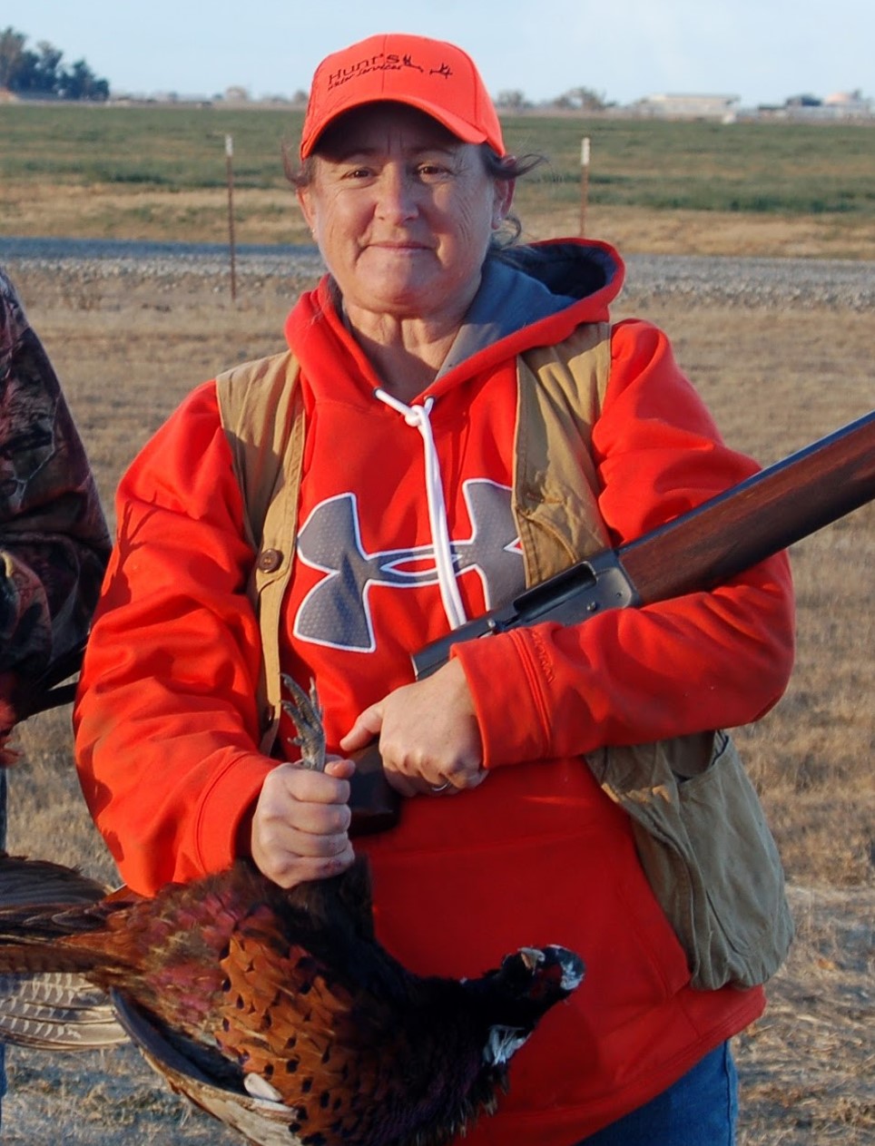 A woman in red baseball cap and hoodie, wearing a khaki vest, holds a rifle and a pheasant which she has shot. Behind her the land is flat and the grass is brown. It appears to be windy.