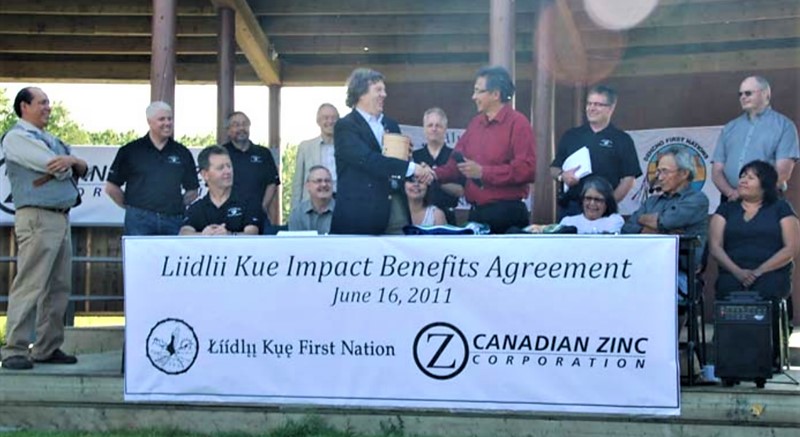 Two people, standing and shaking hands, behind a desk where several people are seated. More people are standing behind them on the covered stage. There is a banner in front of the desk which reads "Liidlii Kue Impact Benefits Agreement - June 16, 2011." The banner has two logos - the one of Liidlii Kue First Nation, and the one of Canadian Zinc Corporation.