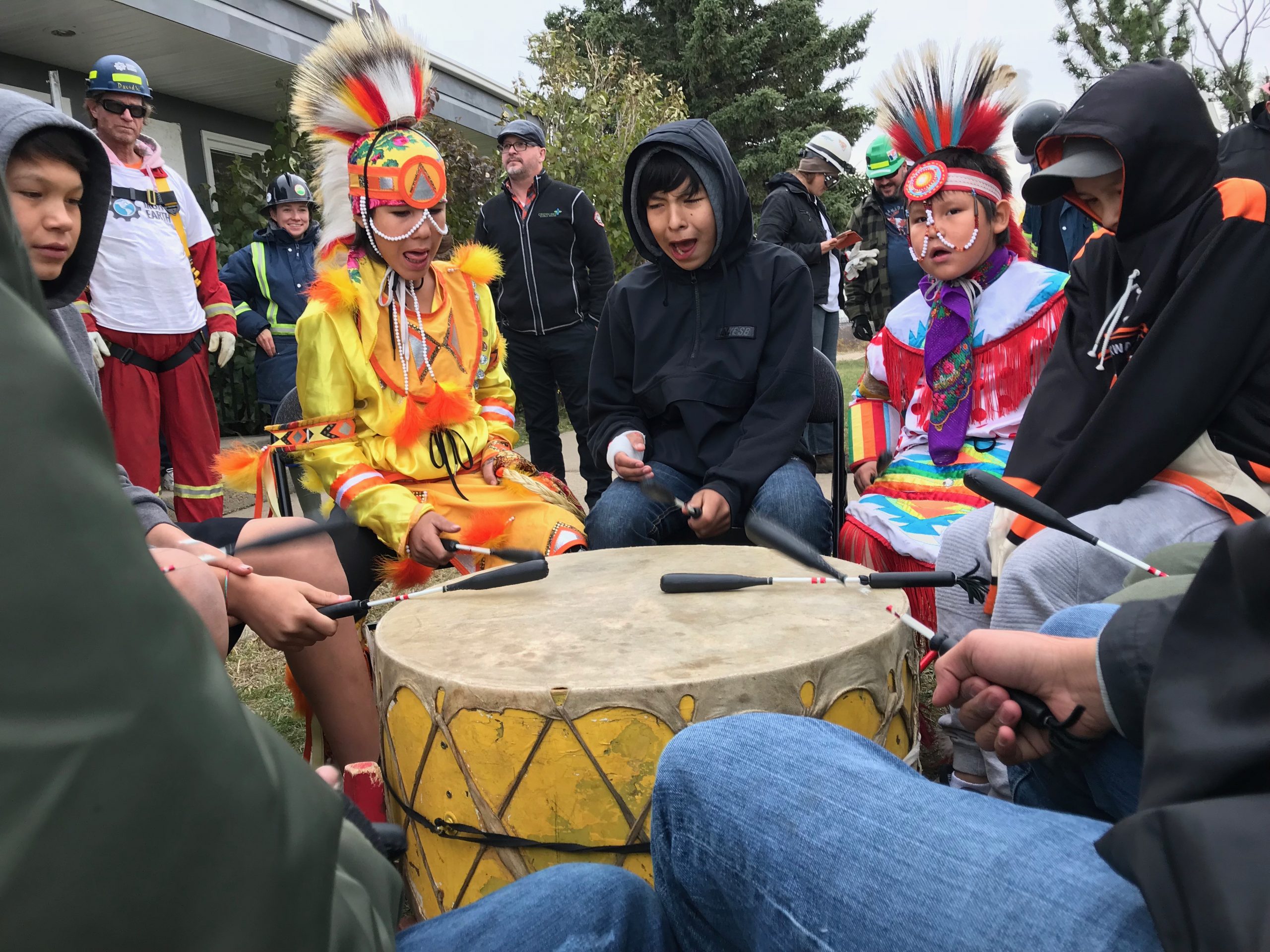 Several boys, approximately 9-13 years of age, some in traditional regalia, seated in a circle outdoors, drumming.