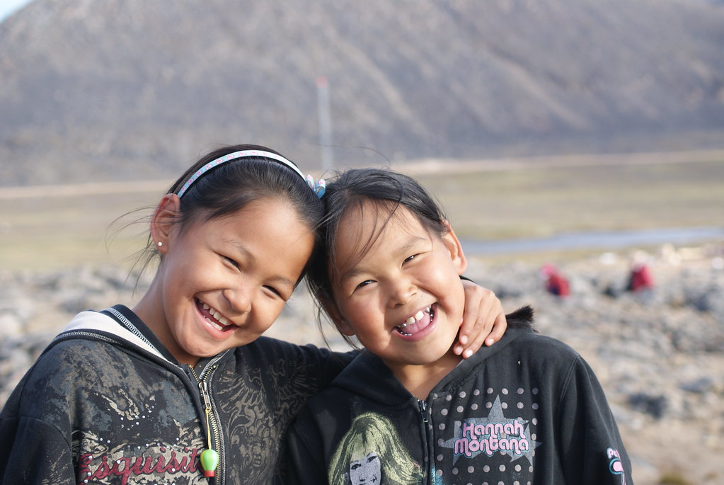 Two smiling Inuit girls, touching heads. One has her hand around the other's neck in a gentle way. Behind them is a rocky area where two blurry figures in red are seated. Beyond that is a grassy lowland with a creek, and beyond that a rocky hill.