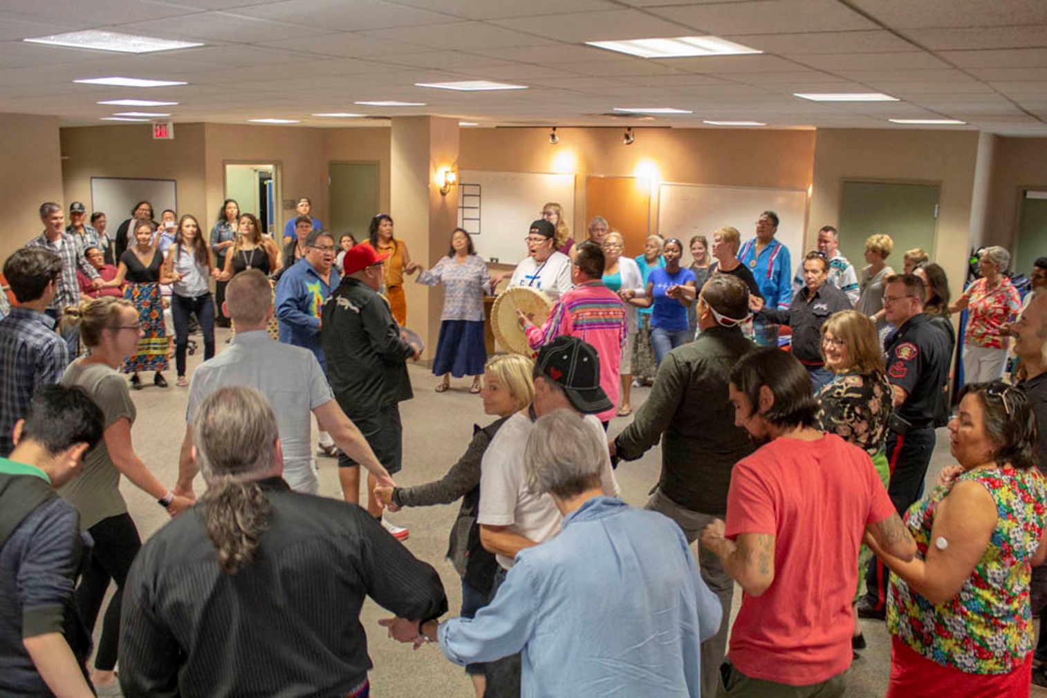 A large carpeted room in a public building, warmly lit, with one or two men holding hand drums in the centre, and two concentric circles of people holding hands around him, ready to dance. Some people are wearing ribbon shirts or skirts.