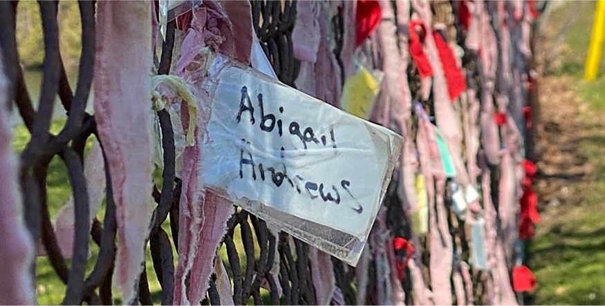 Scraps of fabric, including some red ribbons and name tags, tied to what appears to be an old, rusty chain-link fence. The closest name tag reads "Abigail Andrews".