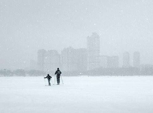 Two figures (adult and child) cross country skiing on snow. Cityscape visible in background.