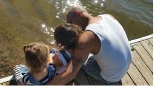 A father sitting on a dock with his arms around his two young children. 