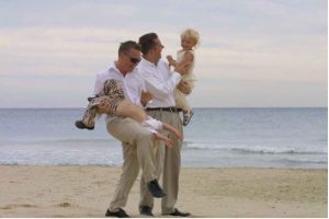 Two men (parents) walking along the beach holding their two young children. 