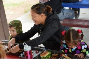 A mother sitting at a pcnic table with her two young children, helping them eat their lunch. 