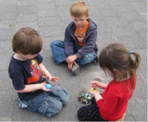 3 children sitting on the pavement playing with toys.