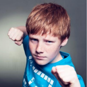 Picture of a young boy with red hair with an aggressive stance, fists up and squinting into the camera.