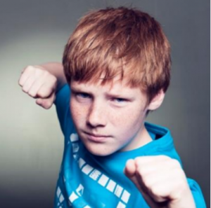 Picture of a young boy with red hair with an aggressive stance, fists up and squinting into the camera. 