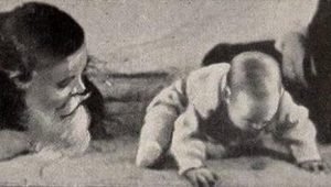 black & white image of baby attempting to crawl with mother laying on the ground watching beside him.