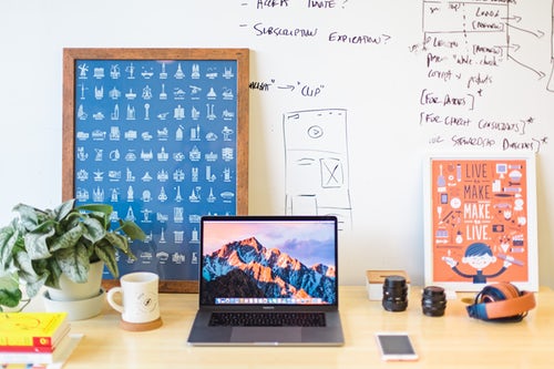 desk with poster of symbols in the background, a computer on the desk and a coffee cup, phone, and other small items well organized