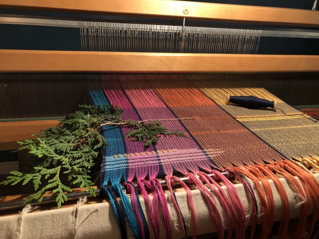a loom, strung with yarn and with a cedar branch resting on the woven cloth