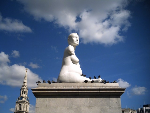 Figure 1-10. A photograph of the white marble sculpture of Alison Lapper. She sits atop a large stone plinth, naked, with her breasts and belly, at seven months pregnant, displayed. Her shortened right leg is outstretched, and her right arm seems to end just below the head of the humerus. The photo is set against a blue sky, with the head of the short-haired figure gazing out the right of the frame, into the sun.